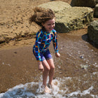 A child in a UV Protective Surf Set Multi Check plays in the shallows at the beach. They are smiling as they splash the water.